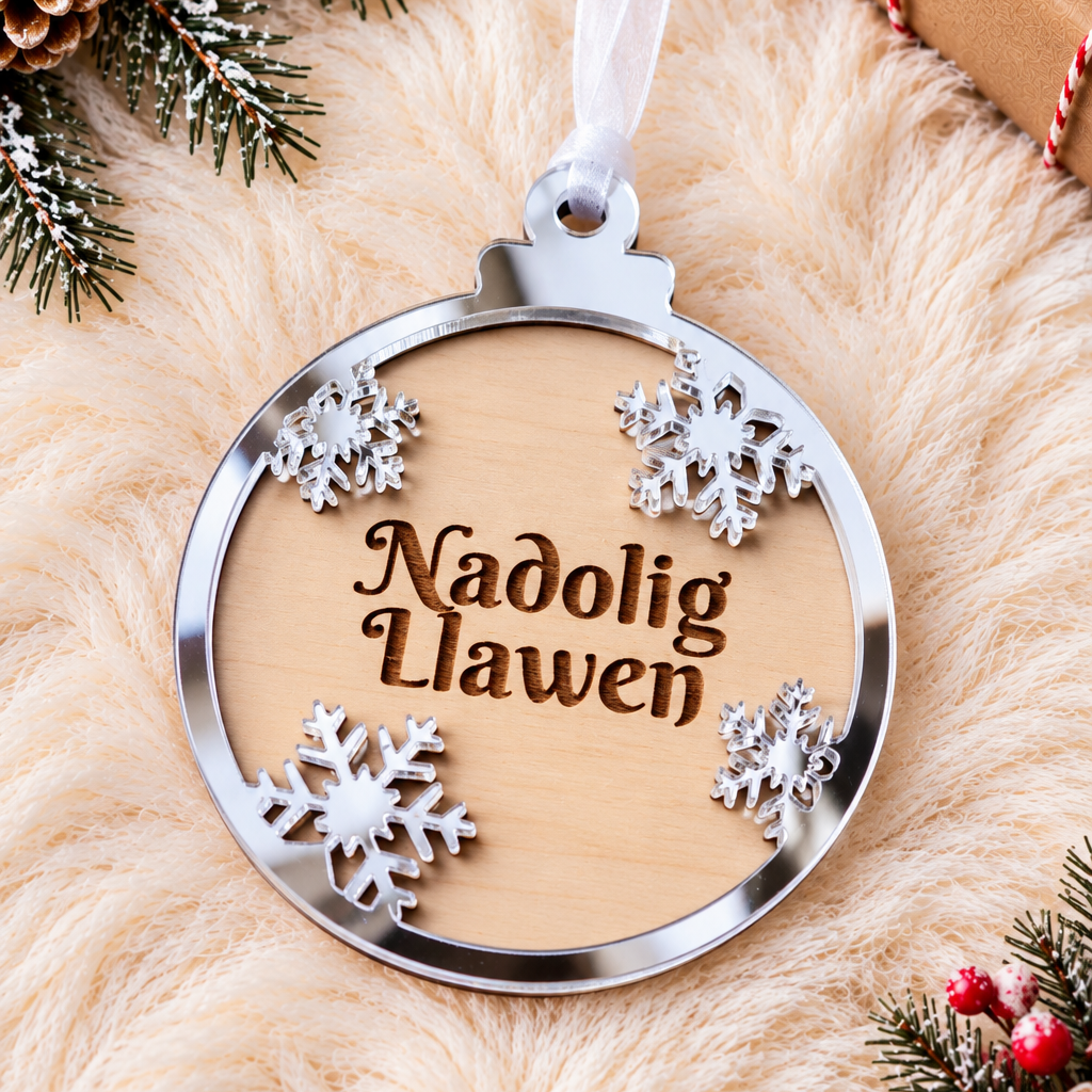 A Welsh Christmas ornament featuring a light wood centre engraved with 'Nadolig Llawen' and surrounded by silver metal snowflakes and a polished silver border, hanging from a white ribbon on a beige faux fur surface with holiday decorations.